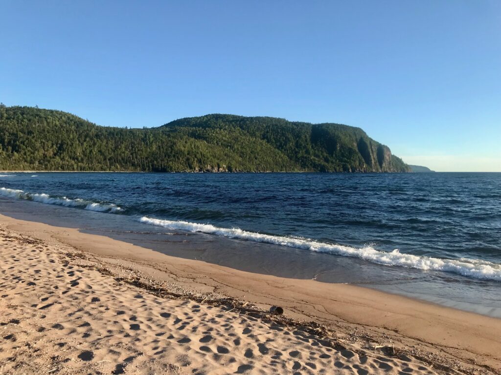 A sandy beach with deep blue water and a forest covered ridge in the background.