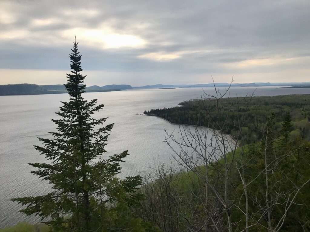 A lake lined with green pine trees and hills and a cloudy sky.